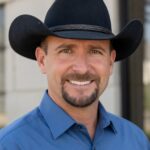 Smiling man in a black cowboy hat and blue shirt, standing outdoors against a neutral background.